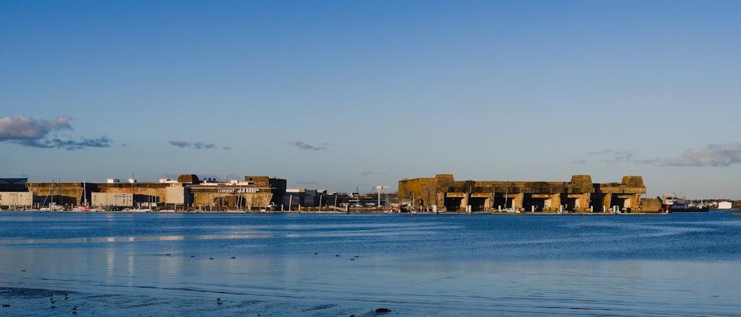 A Distant View of the Old Naval Base in Lorient, France, on a Sunny Afternoon - A large, old naval base sits on the waterfront in Lorient, France. The base is made up of several large, concrete structures and is surrounded by calm water. The sky is blue and clear, and the sun is shining brightly.